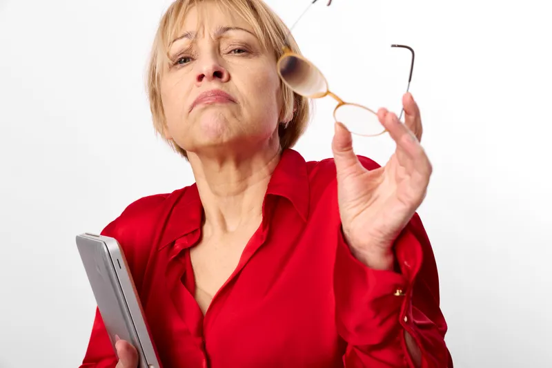 Elderly woman indoors, checking glasses, holding smartphone, concerned expression, vision discomfort, soft light, white background, close up studio portrait