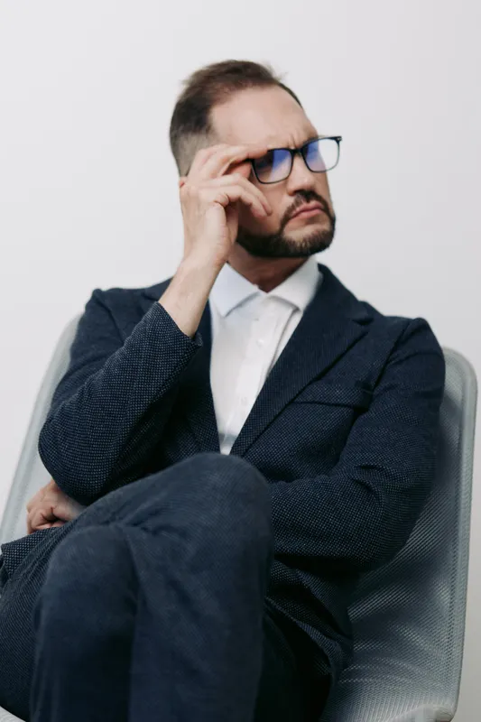 Professional businessman, thoughtful and confident, sits with folded arms and hand on temple, against a solid colored background in a clean studio setting.