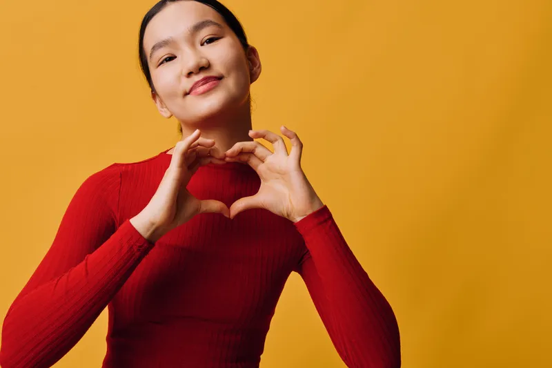 Woman in red sweater making heart shape with hands smiling at camera on yellow background. Portrait of happy Asian female showing love gesture for positive emotion and health.