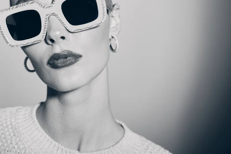 Stylish white woman with fair skin and sunglasses poses in knitted sweater. Fashion portrait with modern accessories and monochrome background.