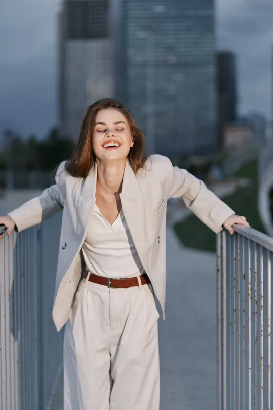 smiling woman in light beige suit laughing outdoors near city buildings, casual fashion style, natural joy and freedom, urban background, spring season