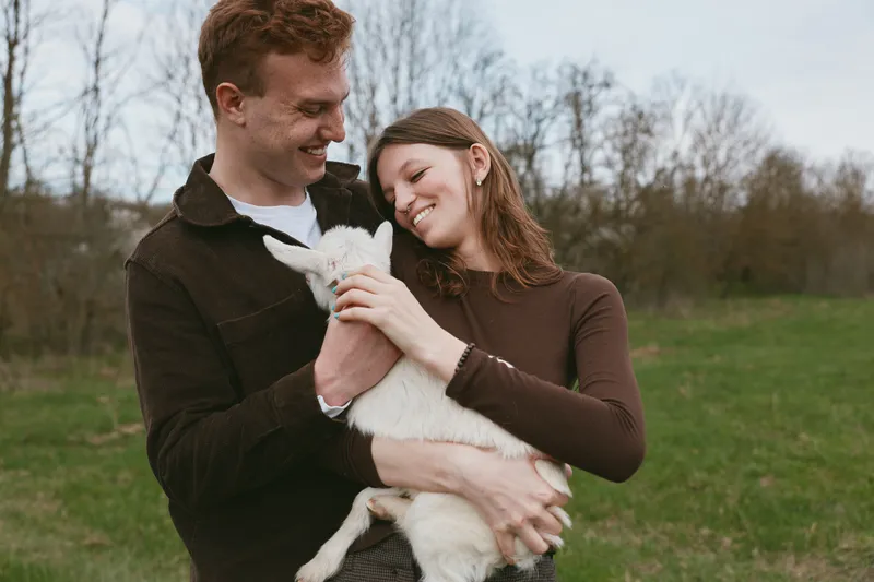 Joyful couple outdoors, holding a fluffy goat kid, warm affection, bright sky, casual attire, grassy field, natural colors, candid moment, rustic charm, celebration of pet companionship
