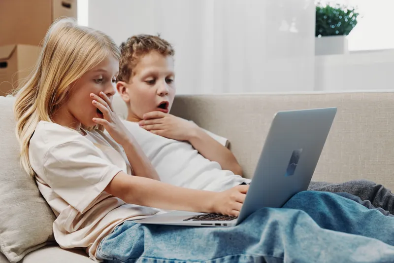 Two surprised children sitting on a couch using a laptop together. The girl and boy show shocked expressions while browsing content in a bright living room environment.