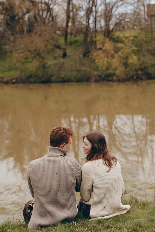 Cheerful couple outdoors by a calm lake, sharing a quiet moment in cozy sweaters, autumn colors reflecting on the water, soft light and breeze, natural setting, relaxed connection, candid lifestyle
