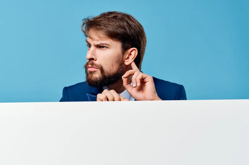 Бородатый мужчина стоит у белого рекламного щита на синем фоне, оставляя место для текста