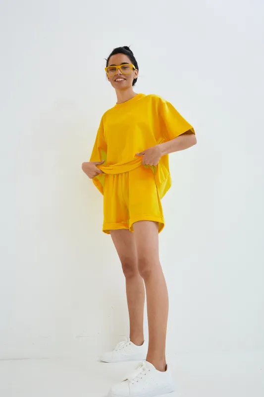 young woman in bright yellow casual outfit and matching sunglasses stands smiling against white background, showcasing relaxed summer fashion and sporty white sneakers.