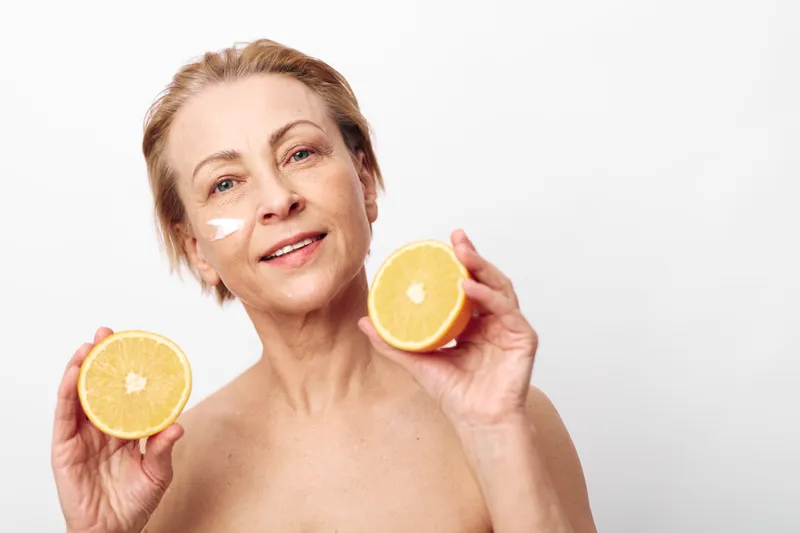 Smiling mature woman with short hair holds two halves of fresh orange, showing natural skincare cream on face. Concept of healthy lifestyle, vitamin skincare, and self-care beauty.