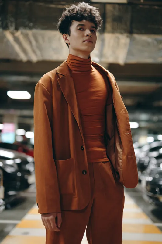 Модный молодой человек в фактурном оранжевом костюме и водолазке позирует в тускло освещенном паркинге
