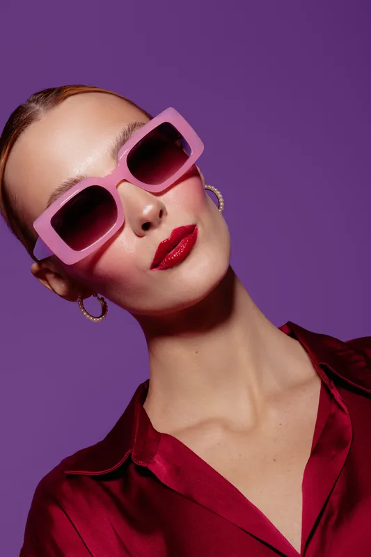 Stylish woman with pink sunglasses and red lipstick poses against purple background wearing red satin blouse and gold hoop earrings. Fashion and beauty concept with confident attitude.