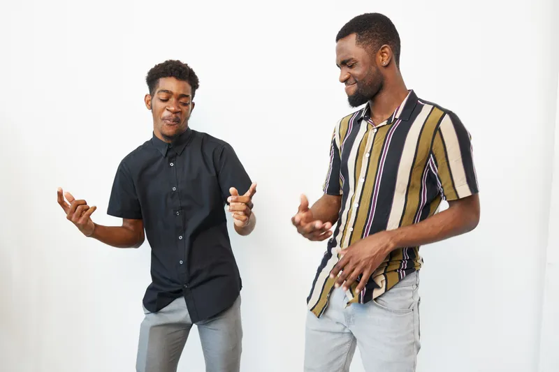 Two smiling African American men dressed in stylish outfits are enjoying a cheerful moment together in a bright, minimalistic setting. Their expressions reflect joy and camaraderie.