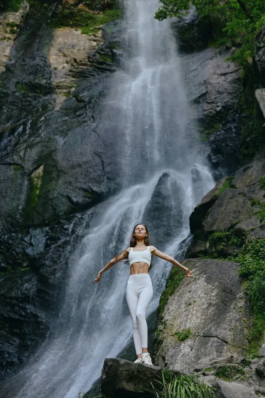 Woman standing near waterfall in white activewear, surrounded by lush green foliage and rocks, expressing freedom and connection with nature in a serene outdoor scene.