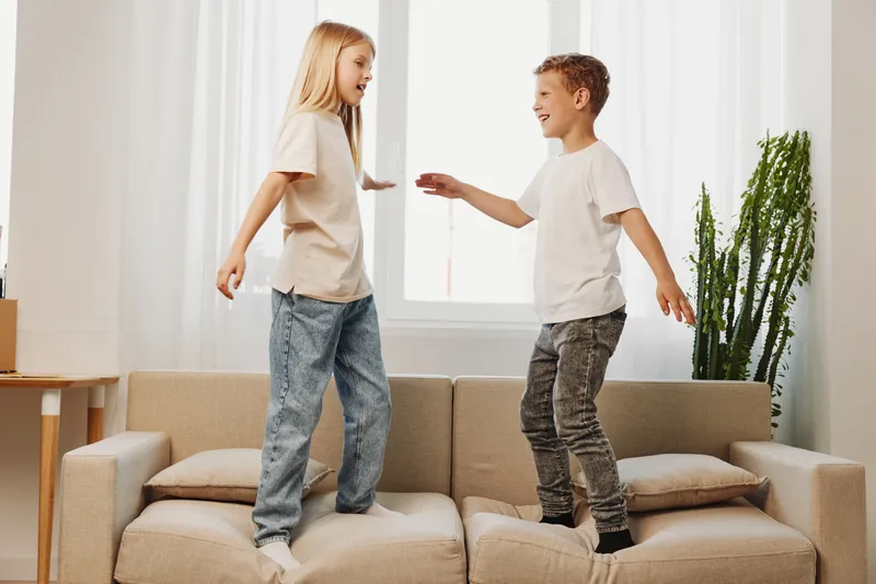 Two happy children jumping on beige sofa in bright living room with large window and green plant, enjoying playful moments together.