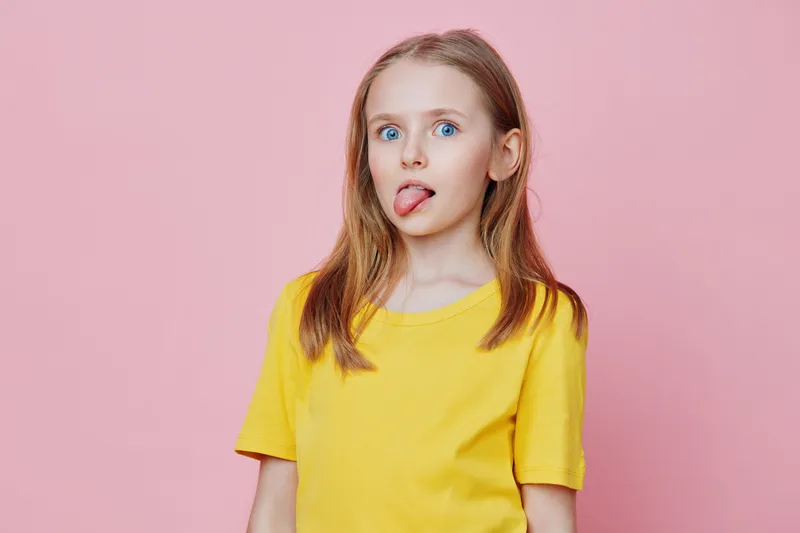 A playful young girl with long blonde hair sticking her tongue out, wearing a bright yellow t shirt, against a soft pink background, capturing a fun and vibrant moment