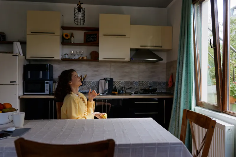 Smiling woman in a yellow top sits at a kitchen table, enjoying a snack. Bright daylight through a window highlights a cozy home dining scene and wholesome lifestyle.