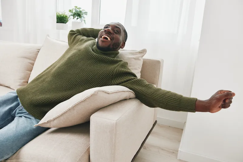 Happy African American man relaxing on a sofa in a cozy sweater, enjoying comfort and tranquility in a bright, modern living room space