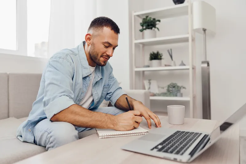 Young man writing in a notebook while sitting on a couch, showcasing creativity and productivity in a cozy, modern workspace environment.