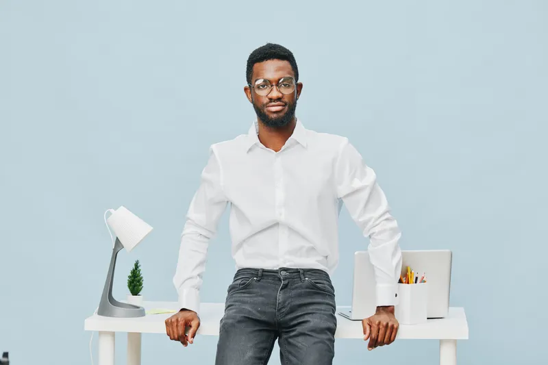 Confident young African American man in white shirt standing behind a desk in modern office space with a laptop and stationery in the background. Business lifestyle concept.