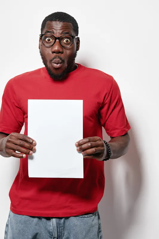African American man with dark skin wearing glasses and red shirt holding blank white paper with surprised expression on face against white background in studio.