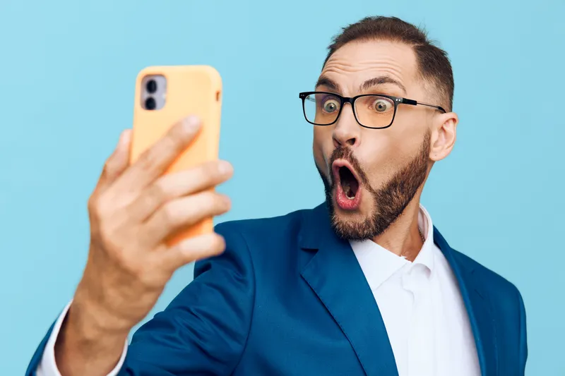 A business professional man on a solid colored background, captured in a candid selfie moment, showing surprise and engagement with the camera.