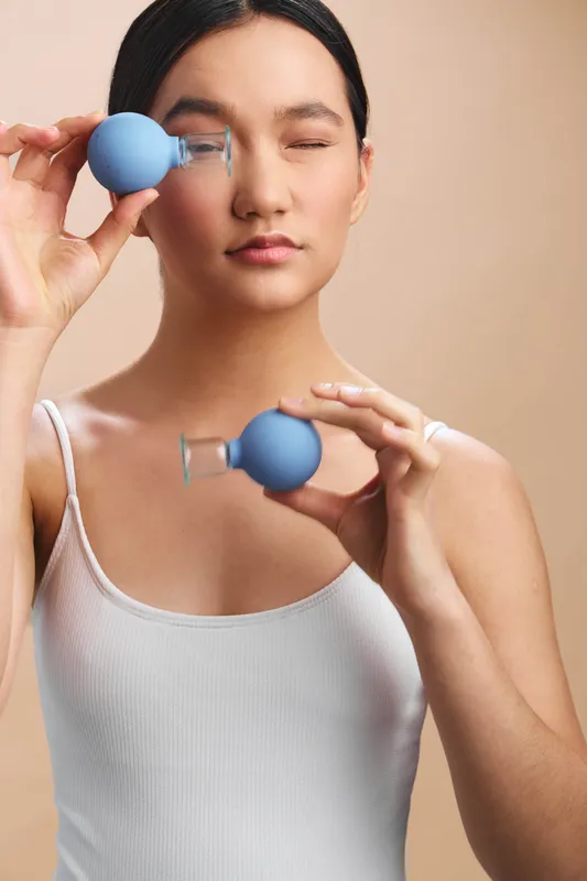 asian woman in a light tank top gently holds blue eye care tools near her eyes with a calm, focused expression, conveying self care and a serene, modern studio atmosphere.