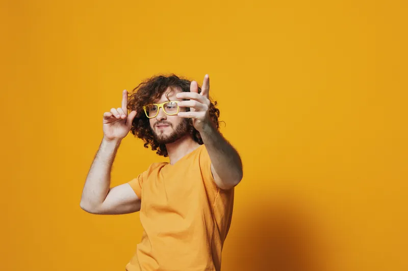 Joyful young man with curly hair in yellow glasses posing against a vibrant yellow background, conveying a sense of creativity and playfulness