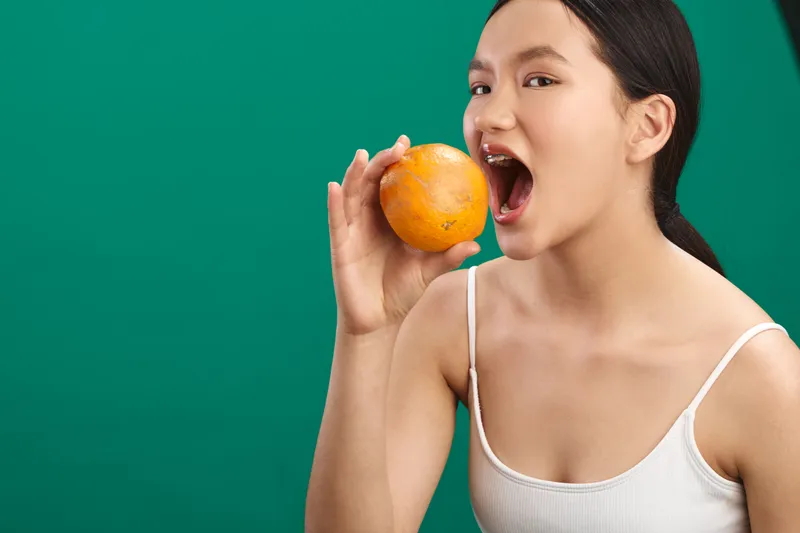 Asian woman enjoys a fresh orange in a bright studio, exuding playful energy and healthy lifestyle vibes as she bites into the fruit with a cheerful, confident smile.