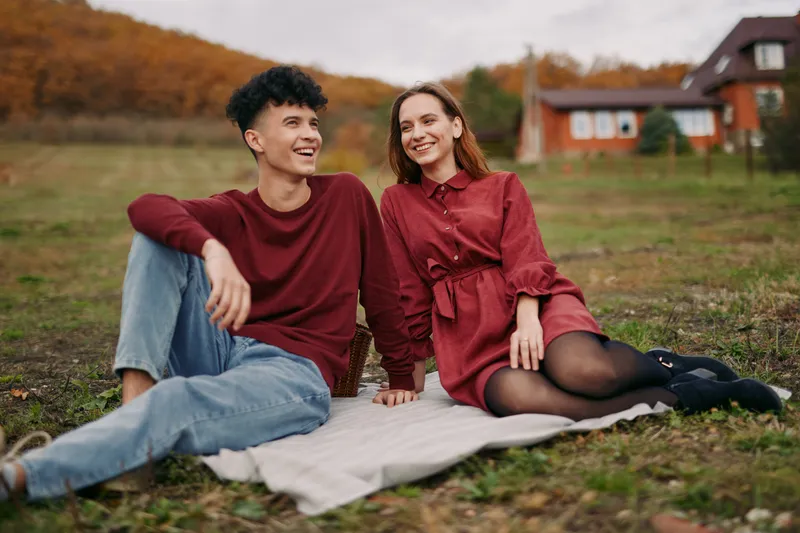 Young couple relaxing on a blanket in a rustic autumn field, sharing genuine smiles and natural interaction that suggest authentic moments and credibility in everyday outdoor life.