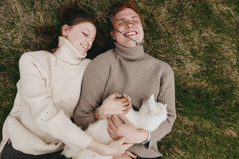 Cheerful couple outdoors, lying on grass, cozy sweaters, smiling softly with a small goat kid beside them, relaxed mood, natural colors, warm light, lifestyle moment