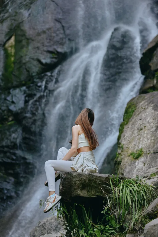 woman sitting near waterfall in white activewear, long hair flowing, natural environment with rocks and greenery, serene outdoor scene with motion water.