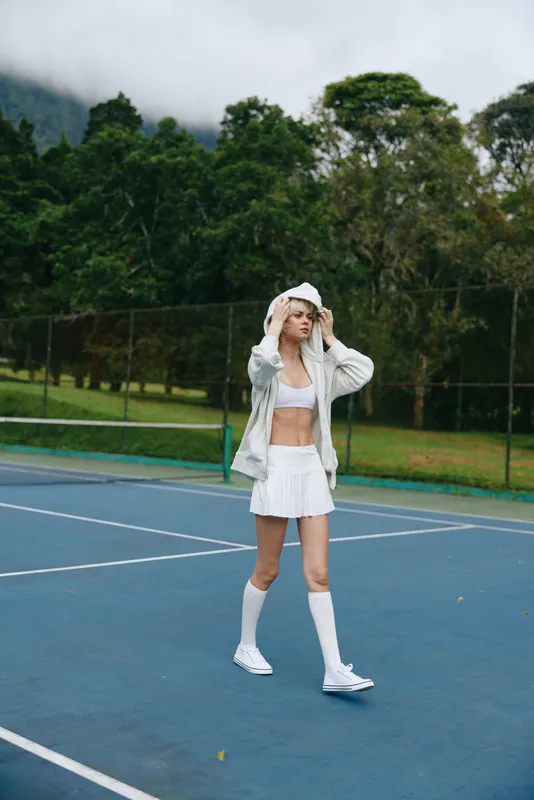 Athletic young woman in sporty outfit walking confidently on tennis court, wearing hoodie and skirt, showcasing athleticism and style in a lush green setting