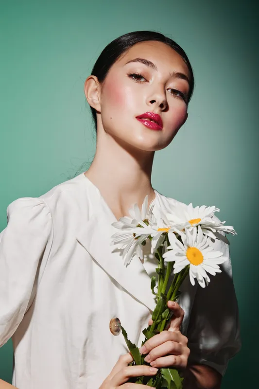 Young woman with bright makeup holds white daisies. She wears a white blouse against a soft green background. Concept of fashion beauty creative portrait, freshness and elegance.