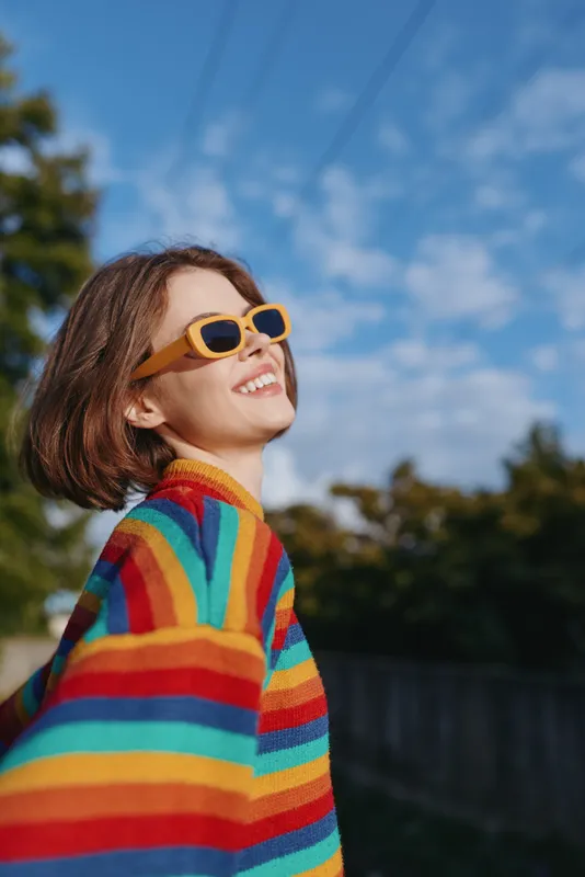 Young adult woman smiling in a rainbow sweater and yellow sunglasses outdoors under blue sky, short bob hair, joyful expression, casual fashion portrait enjoying sunny urban nature.
