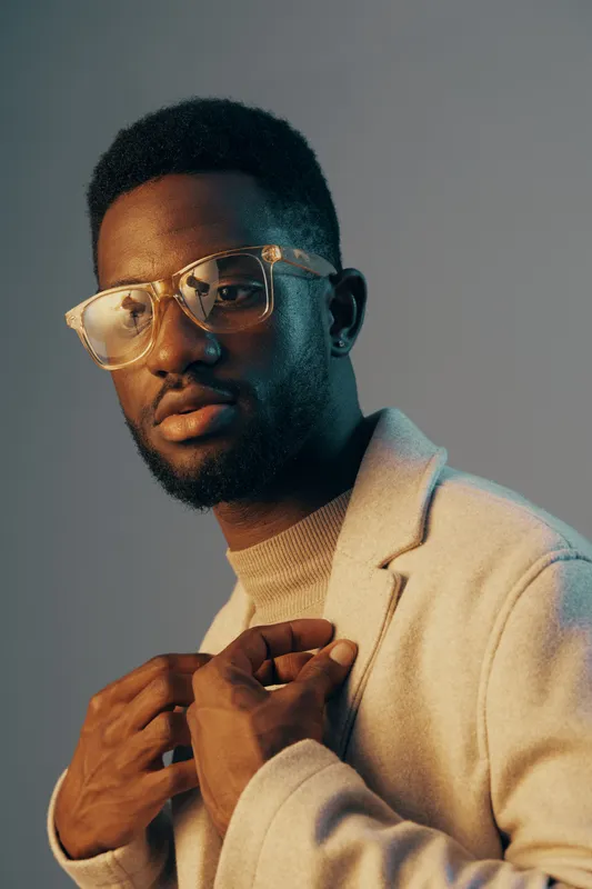 Stylish young man with glasses posing elegantly in a beige coat, showcasing confidence and fashion forwardness against a soft blue background