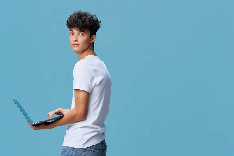 Young man using laptop wearing casual white t-shirt and jeans, standing against blue background with focused expression and modern hairstyle.