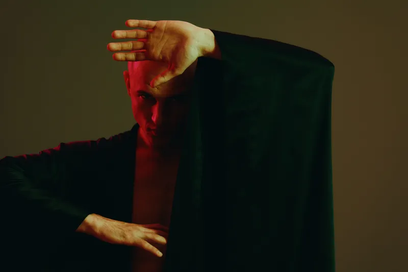 Striking man in black attire, showcasing a dramatic pose with a hand raised, emphasizing strength and emotion in a dimly lit scene.