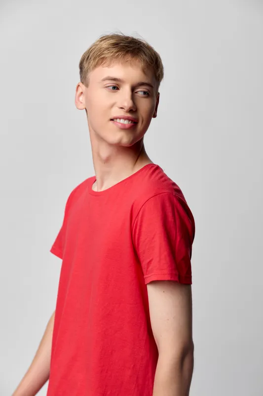 Red shirt, smiling boy in casual fashion stands in studio photography setting with bright background, relaxed pose, and cheerful mood for a youth oriented, teen friendly casual look