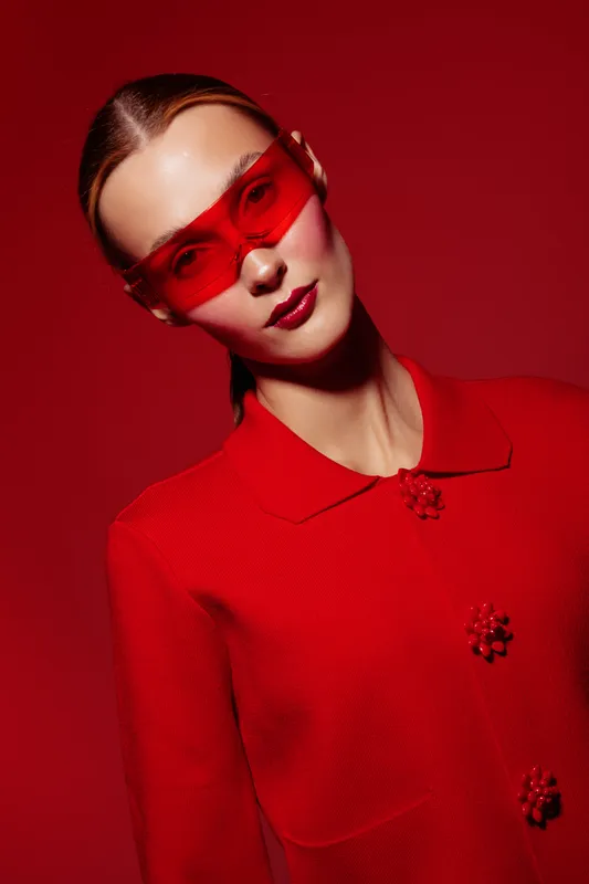 Stylish woman in red coat and red sunglasses poses against red background. Fashion portrait with bold color contrast and modern accessories. Concept of style, elegance, and confidence.