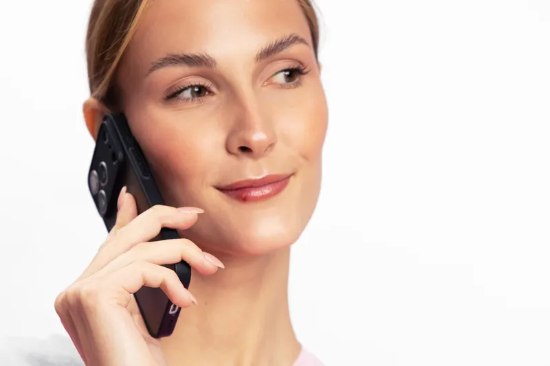 Young woman with clear skin holding smartphone near ear, smiling slightly, looking away. Concept for communication, technology, mobile phone, and positive emotions in studio.