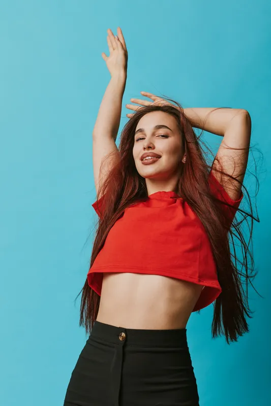 Young woman with long hair wearing red crop top and black pants posing with hands raised against blue background smiling confidently in studio portrait fashion photo.