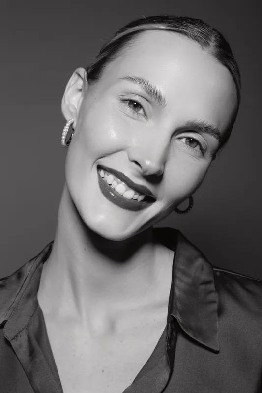Smiling Caucasian woman with pale skin and slicked-back hair wears hoop earrings and a satin blouse. Studio portrait in black and white lighting.