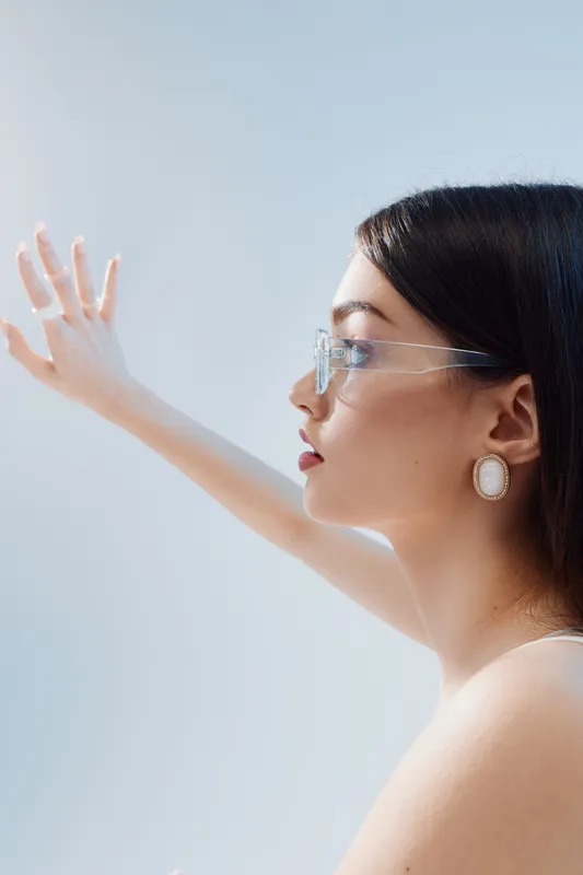 Profile of a young woman wearing transparent glasses and elegant earrings reaching out with her hand. Soft natural light highlights her beauty and fashion style.