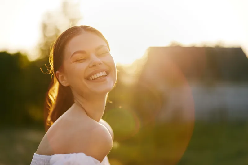 Happy young woman laughing in sunlight, wearing a white off shoulder dress, with a warm golden background, conveying joy and positivity in a summer setting