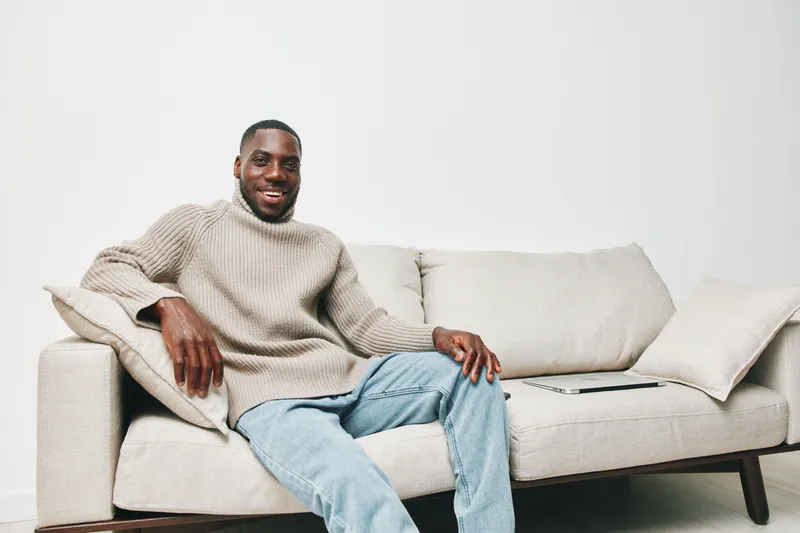 Happy young Black man sitting on a sofa in cozy sweater, smiling and enjoying a relaxed moment at home, promoting comfort and lifestyle concepts