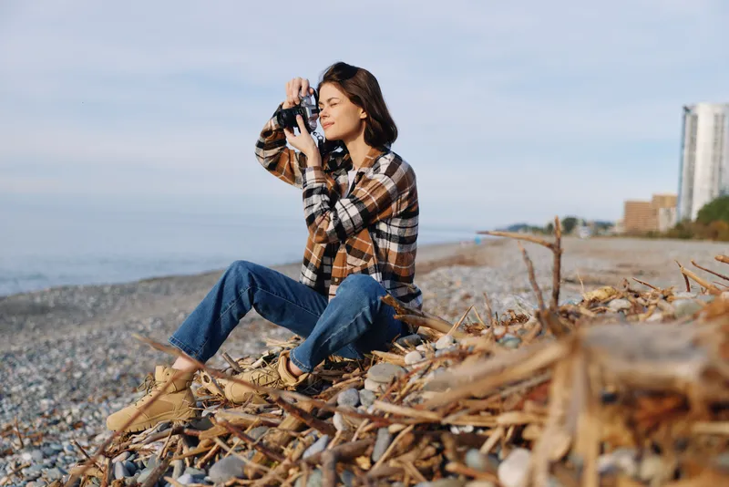 Woman photographer on a rocky beach photographs with a handheld camera, wearing a plaid shirt and jeans, driftwood and calm sea creating a relaxed outdoor scene for nature lovers and travel