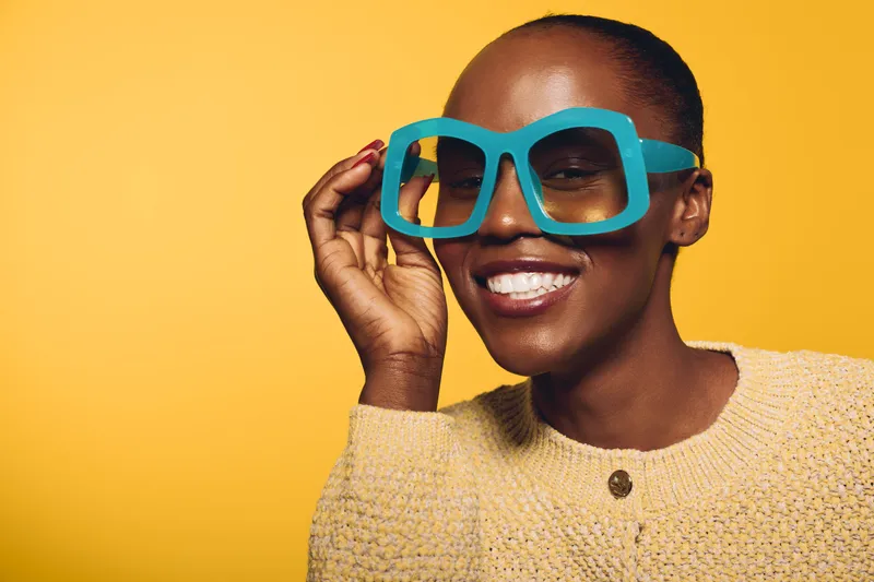 Smiling dark skinned African woman with short hair wearing oversized blue glasses and beige sweater poses against yellow background. Concept of fashion, joy, and modern style.