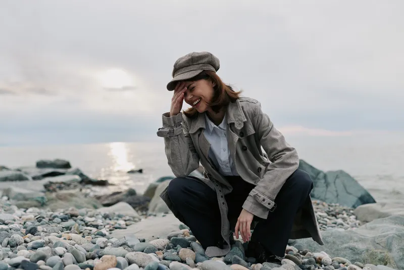 Young woman in stylish coat and hat squats on rocky beach near ocean, smiling and covering face with hand. Overcast sky and calm sea create peaceful autumn mood outdoors.