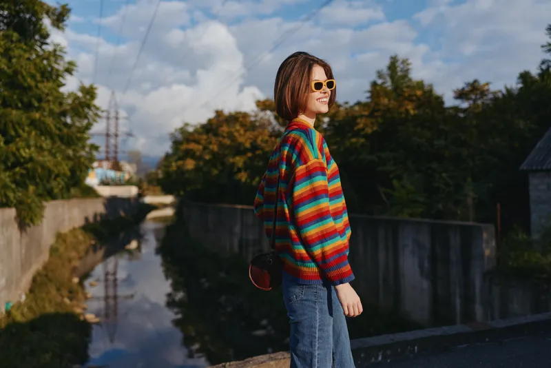 Woman in colorful striped sweater and sunglasses with braided hair and jeans standing by urban canal smiling, casual travel lifestyle portrait in small town outdoors.