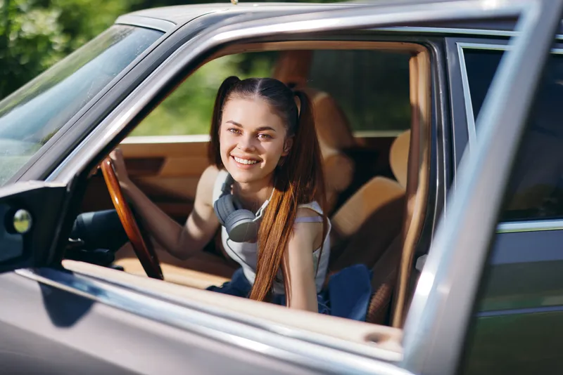 Cheerful young woman with headphones smiling from the driver's seat of a vintage car on a sunny day, perfect for concepts of travel and freedom.