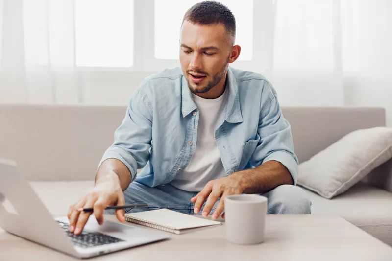 Focused man working on a laptop at home, enjoying a productive moment. He is dressed casually while taking notes and sipping coffee, exuding creativity and concentration.