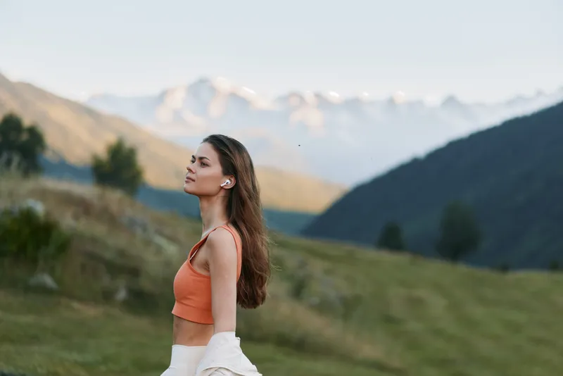 A calm young woman is seen standing in a scenic mountain landscape during golden hour. She wears an orange crop top and light pants, gazing toward distant peaks, with a peaceful and expansive outdoor